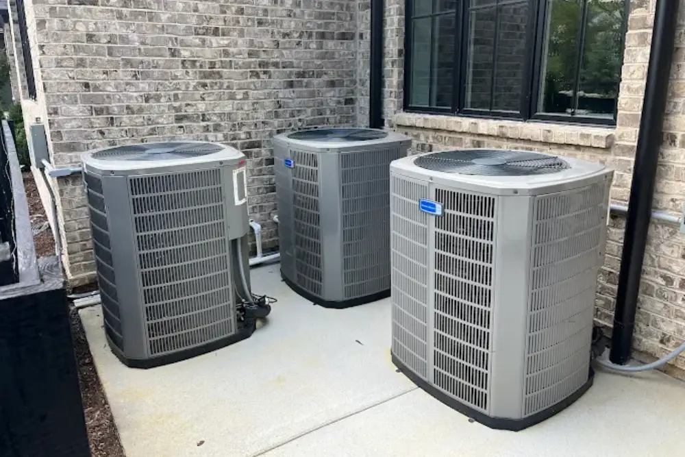 Three gray air conditioning units sit on a concrete slab beside a brick wall, with windows and pipes visible in the background